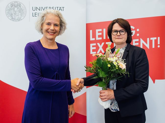 Rektorin Obergfell steht links, Professorin Latzko rechts, beide geben sich die rechte Hand. Frau Latzko hält in der linken Hand einen bunten Blumenstrauß. 