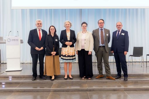 Eine Gruppe von sechs Personen vor einem hellen Vorhang, nebeneinander stehend.