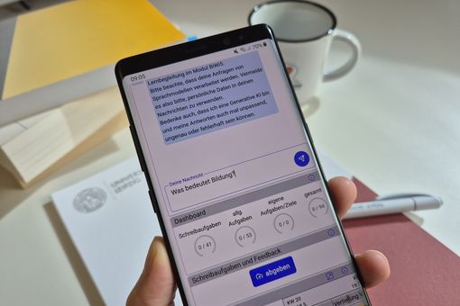 Hand hält ein Smartphone, auf dem der Chatbot zu sehen ist. Die eingetippte Frage lautet: "Was bedeutet Bildung?"