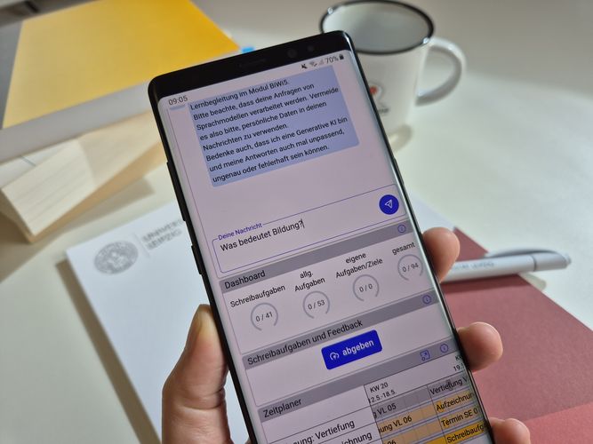 Hand hält ein Smartphone, auf dem der Chatbot zu sehen ist. Die eingetippte Frage lautet: "Was bedeutet Bildung?"
