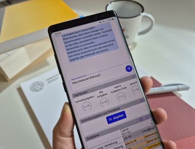 Hand hält ein Smartphone, auf dem der Chatbot zu sehen ist. Die eingetippte Frage lautet: "Was bedeutet Bildung?"