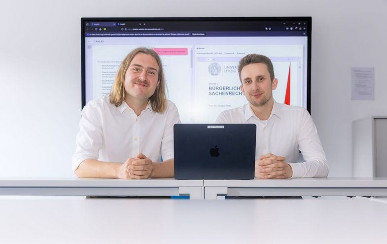Zwei junge Männer sitzen an einem Tisch vor einem Großbildschirm, auf dem eine Internetseite zu sehen ist. 