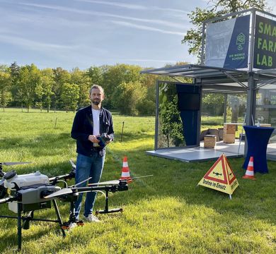Wirtschaftsinformatiker Martin Schieck überprüft die Drohne nach ihrem Flug übers Versuchsfeld. Foto: Diana Smikalla Auf dem Bild ist Wirtschaftsinformatiker Martin Schieck auf einer Wiese neben einer Drohne zu sehen. Neben ihm das Smart Farming Lab.ersuchsfeld. Foto: Diana Smikalla