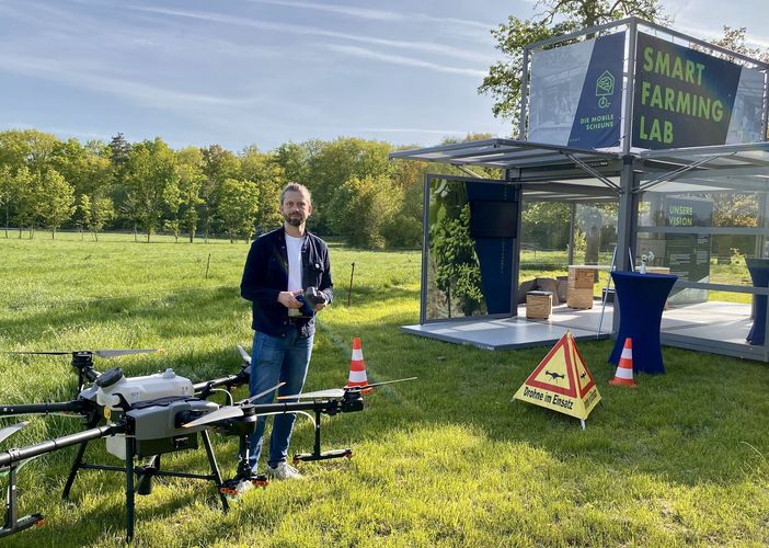 Auf dem Bild ist Wirtschaftsinformatiker Martin Schieck auf einer Wiese neben einer Drohne zu sehen. Neben ihm das Smart Farming Lab.ersuchsfeld. Foto: Diana Smikalla