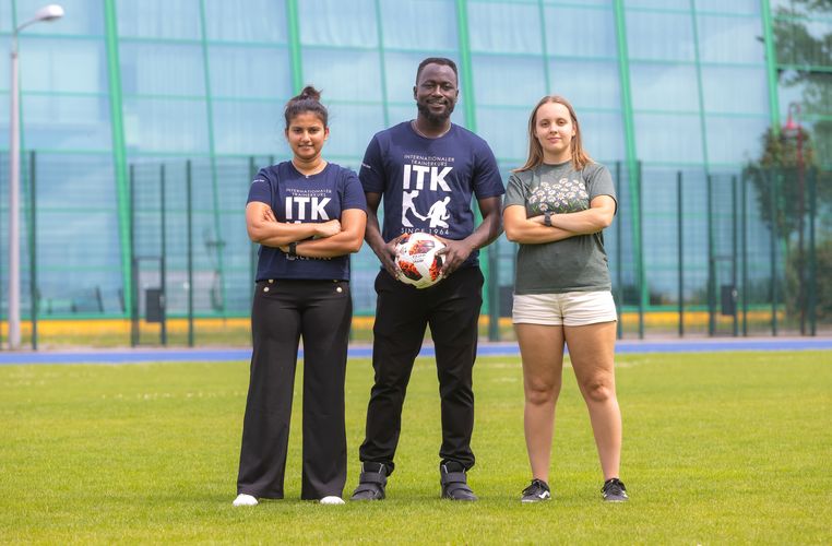 Auf dem Bild sind Yamuna aus Sri Lanka (links), Brian aus Uganda und Mariana aus Brasilien auf der Wiese mit einem Fußball zu sehen.