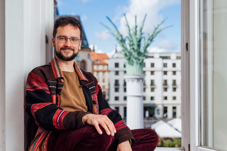 Zu sehen ist Dominik Becher im Fenster seines Büros, im Hintergrund ist der Nikolaikirchhof zu erkennen.