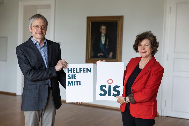 Hans-Bert Rademacher und Beate Schücking halten jeweils ein Schild, auf dem "Helfen Sie mit" bzw. das Logo des Vereins SIS zu sehen ist.