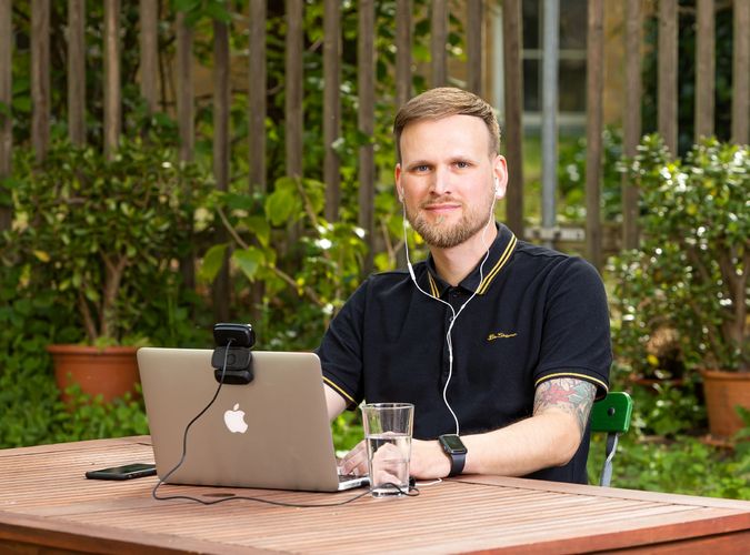 Christoph Bülau sitzt an einem Tisch im Freien. Vor ihm steht ein aufgeklappter Laptop mit aufgesteckter Webcam. Im Hintergrund ist ein Garten zu erkennen.