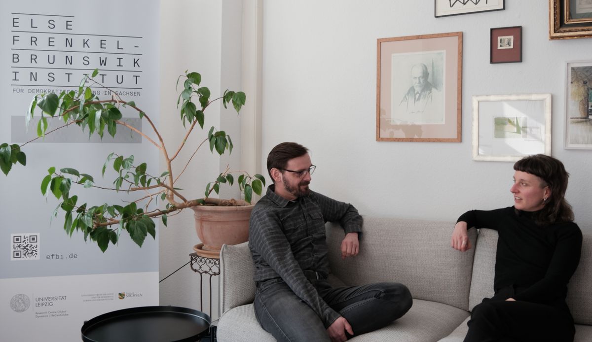 zur Vergrößerungsansicht des Bildes: Zwei Menschen sitzen auf einem Sofa und unterhalten sich