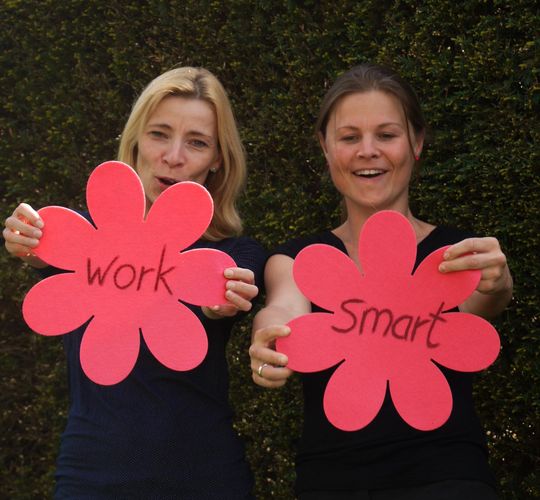 Manuela Süß und Yvonn Semek halten jeweils ein rotes Schild in Blütenform, auf dem die Wörter Work und Smart stehen