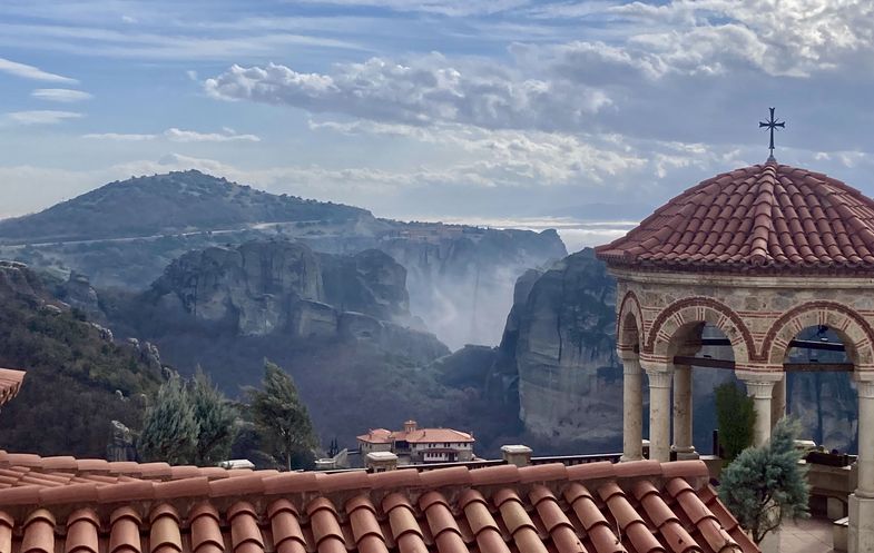 Zu sehen ist eine bergige, felsige und wolkenverhangene Landschaft. Im Vordergrund zu sehen ist ein Kloster.