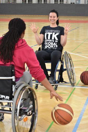 Zwei Rollstuhl-Basketballerinnen mit Ball in einer Sporthalle.