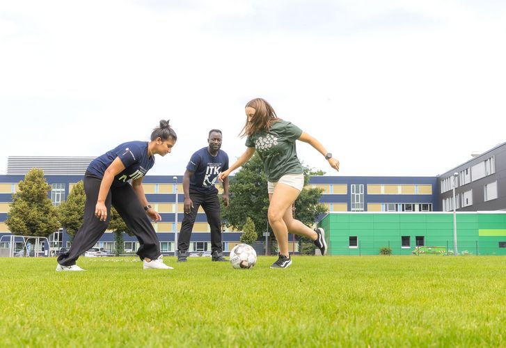 Auf dem Bild sind beim Fußballspielen auf einer grünen Wiese zu sehen: die ITK-Kursteilnehmer Yamuna (links), Brian und Mariana. 