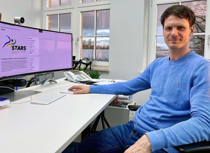 Ein Mann sitzt an einem Schreibtisch, auf dem ein großer Bildschirm steht, mit der Webseite eines Projekts namens 'STARS'. Im Hintergrund sind große Fenster mit Blick auf eine winterliche Landschaft zu sehen.