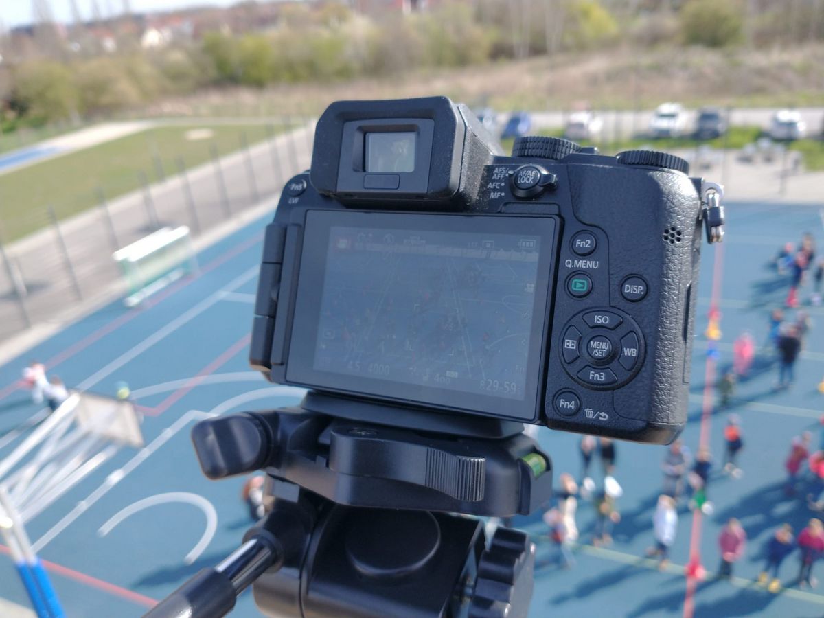 zur Vergrößerungsansicht des Bildes: Zu sehen ist eine Kamera auf dem Dach der Schule, die den Sportplatz filmt, auf dem die Grundschüler:innen eine Choreografie einstudieren.