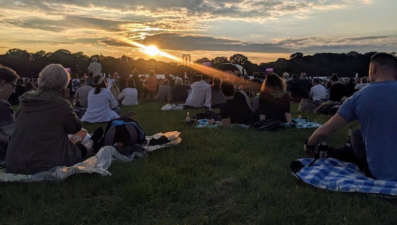 Zu sehen ist eine Bühne für das Open-Air-Konzert "Klassik Airleben" auf einer Wiese im Rosenthal., darüber geht die Sonne langsam unter, davor sitzen viele Menschen auf Picknick-Decken.