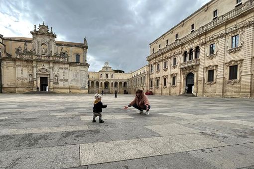 Ein Kleinkind läuft auf einem weitläufigen Platz seiner Mutter entgegen, die in der Hocke ist und ihre Arme dem Kind entgegenstreckt. Der Platz ist umsäumt von historischen Gebäuden. Im Hintergrund sieht man vereinzelt Menschen. 