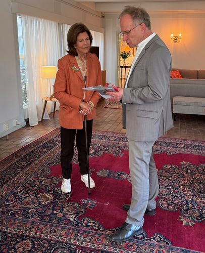 Königin Silvia zu Schweden steht, auf einer Gehhilfe gestützt, neben Prof. Dr. Peter Zimmerling im Gesrpäch. Zimmerling präsentiert ihr ein Geschenk.