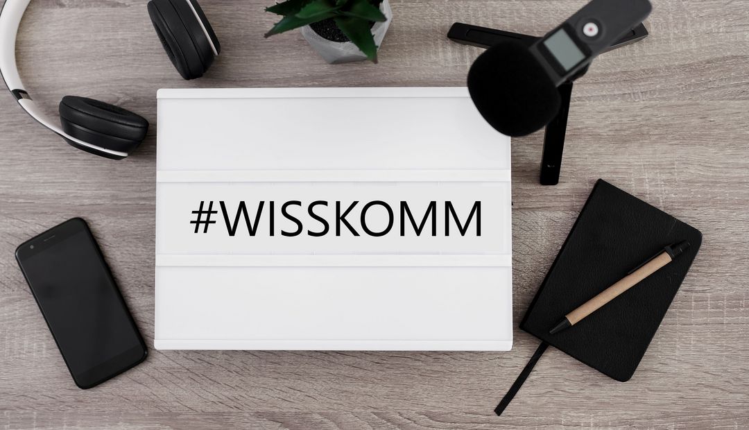 Zu sehen ist ein Schild mit der Aufschrift #WISSKOMM, darum herum liegen ein Notizblock, ein Mikrofon, ein Smartphone und ein Kopfhörer