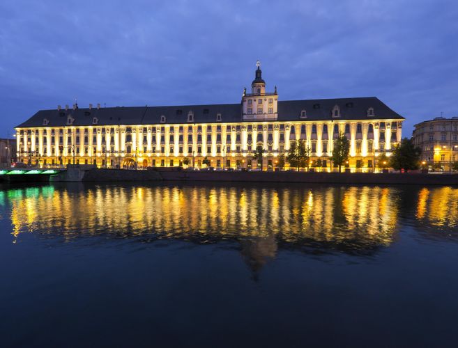 Zu sehen ist die Fassade des barocken und langgezogenen Hauptgebäudes der Universität Breslau, angeleuchtet in der Abenddämmerung. Es spiegelt sich in einem davorliegenden See.