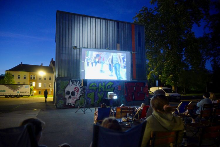 Menschen sitzen in Liegestühlen und sehen im Abendlicht eine Kinovorstellung auf einem Stadtplatz