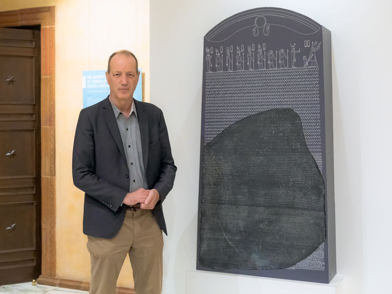 Auf dem Bild ist Dr. Dietrich Raue im Ägyptischen Museum vor einer Kopie des berühmten Rosetta-Steins zu sehen.