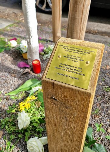 Zu sehen ist das Messingschild, das vor dem Baum angebracht ist, mit der Aufschrift: "Im Gedenken an den Universitätsprofessor Oliver von Knebel Doeberitz. Institut für Anglistik". 